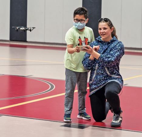 The photo shows a young Alaska Native student flying a tiny drone with help from a DRONES project staff member. They are inside the gymnasium at the Andrew K. Demoski School in Nulato, Alaska.