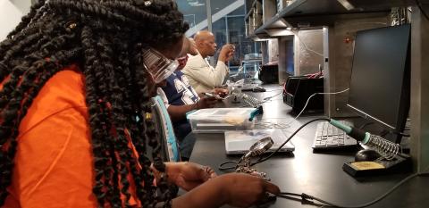 1 woman and 2 men with safety goggles engaged in soldering a circuit board in an electronics lab setting 