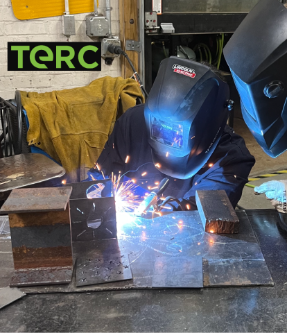 Photo of young woman wearing a welding helmet welding together pieces of steel. A mentor wearing a welding helmet leans in to watch.