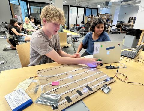 Two teen interns work together using a laptop and an arduino board to program LEDs to light up in a specific pattern. In the background, other teen interns work on their projects.