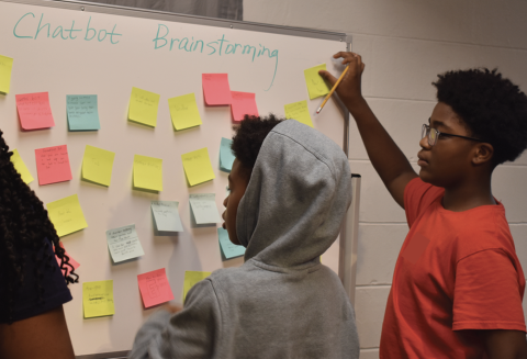 Two young Black students looking at a whiteboard covered with "Chatbot Brainstorming" written at the top. sticky notes, with 